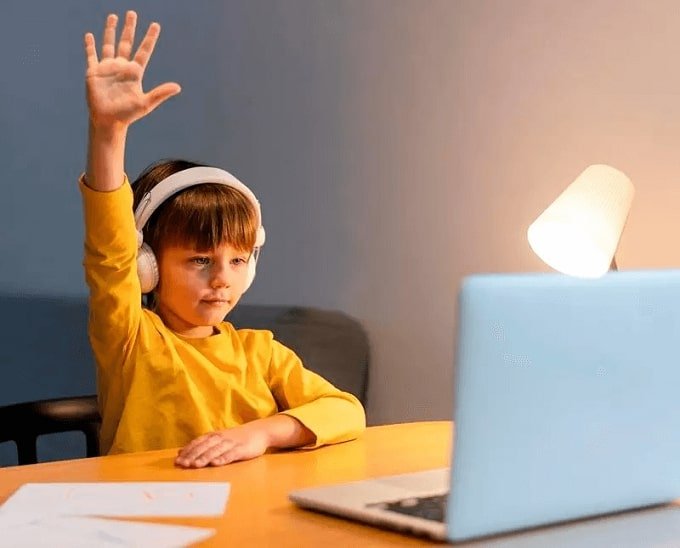 Un enfant avec une chemise jaune et des écouteurs qui lève la main pendant un cours en ligne.